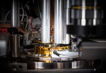 Die Probe, ein zylinderförmiger Ausschnitt aus einem handelsüblichen Computerchip, wird von der goldfarbenen Nadel in der Mitte des Bildes gehalten. Mit einem Durchmesser von weniger als 0,000 005 Meter (etwa 20 Mal kleiner als die Breite eines menschlichen Haares) wurde sie mit einem fokussierten Ionenstrahl aus dem Chip herausgeschnitten und auf dem Stift platziert. © Paul Scherrer Institut PSI/Mahir Dzambegovic