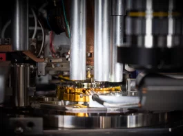 Die Probe, ein zylinderförmiger Ausschnitt aus einem handelsüblichen Computerchip, wird von der goldfarbenen Nadel in der Mitte des Bildes gehalten. Mit einem Durchmesser von weniger als 0,000 005 Meter (etwa 20 Mal kleiner als die Breite eines menschlichen Haares) wurde sie mit einem fokussierten Ionenstrahl aus dem Chip herausgeschnitten und auf dem Stift platziert. © Paul Scherrer Institut PSI/Mahir Dzambegovic