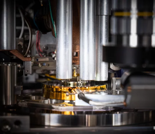 Die Probe, ein zylinderförmiger Ausschnitt aus einem handelsüblichen Computerchip, wird von der goldfarbenen Nadel in der Mitte des Bildes gehalten. Mit einem Durchmesser von weniger als 0,000 005 Meter (etwa 20 Mal kleiner als die Breite eines menschlichen Haares) wurde sie mit einem fokussierten Ionenstrahl aus dem Chip herausgeschnitten und auf dem Stift platziert. © Paul Scherrer Institut PSI/Mahir Dzambegovic