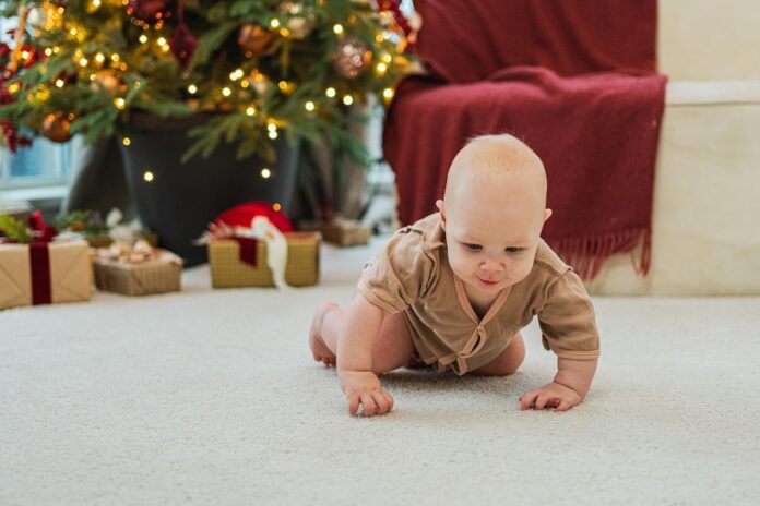 Frohe Weihnachten Xmas. Glücklich neugeborenes Baby Kind krabbeln in der Nähe von Weihnachtsbaum im Wohnzimmer zu Hause. Kleines Mädchen Kind lächelnd im Zimmer mit Weihnachtsschmuck. Happy Baby in ersten Weihnachtsabend.