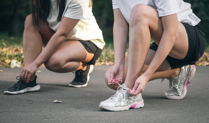 Two young women tie their shoelaces before jogging in the park.